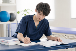 リハビリテーション室のベッドの横で、参考書を広げながら真剣な表情でメモを取る若い男性